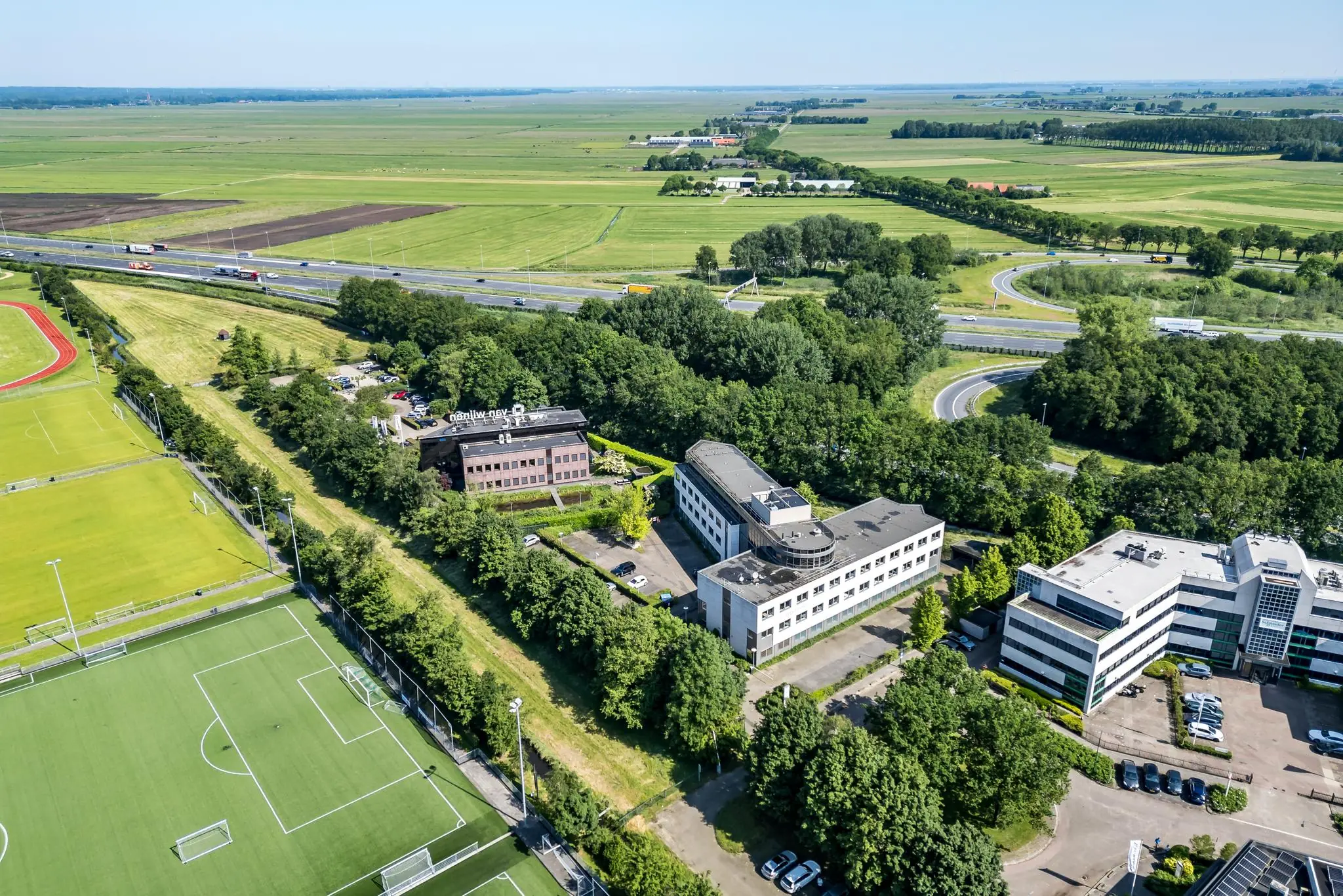 Luchtfoto van kantoren aan de Baarnsche Dijk naast sportvelden en de snelweg, omringd door groen en weilanden.